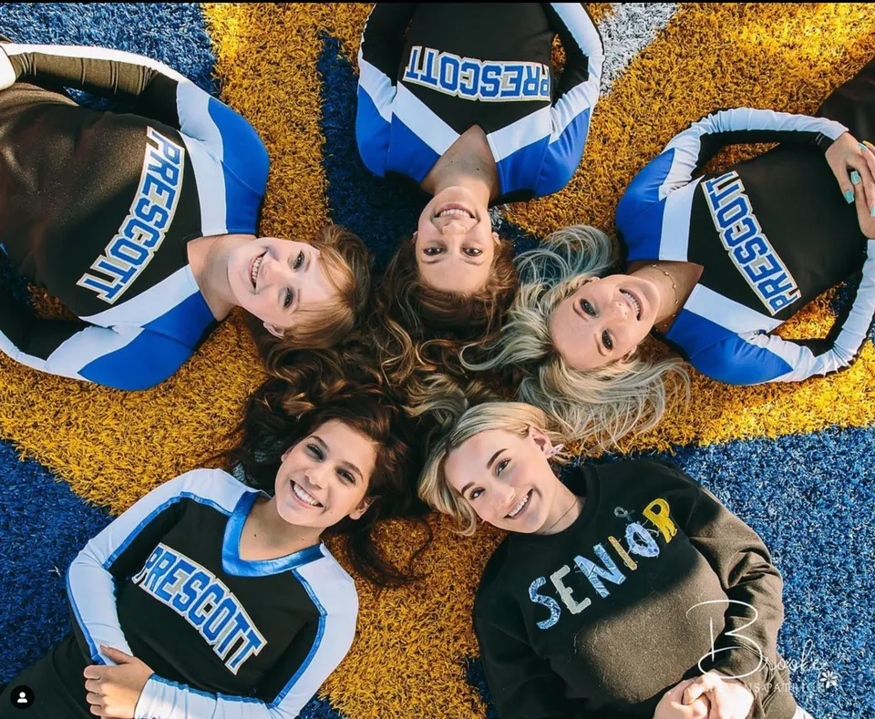 Giovani ragazze in uniformi da cheerleader sono sdraiate sorridenti sul pavimento davanti a uno sfondo colorato nel campus della Prescott High School.