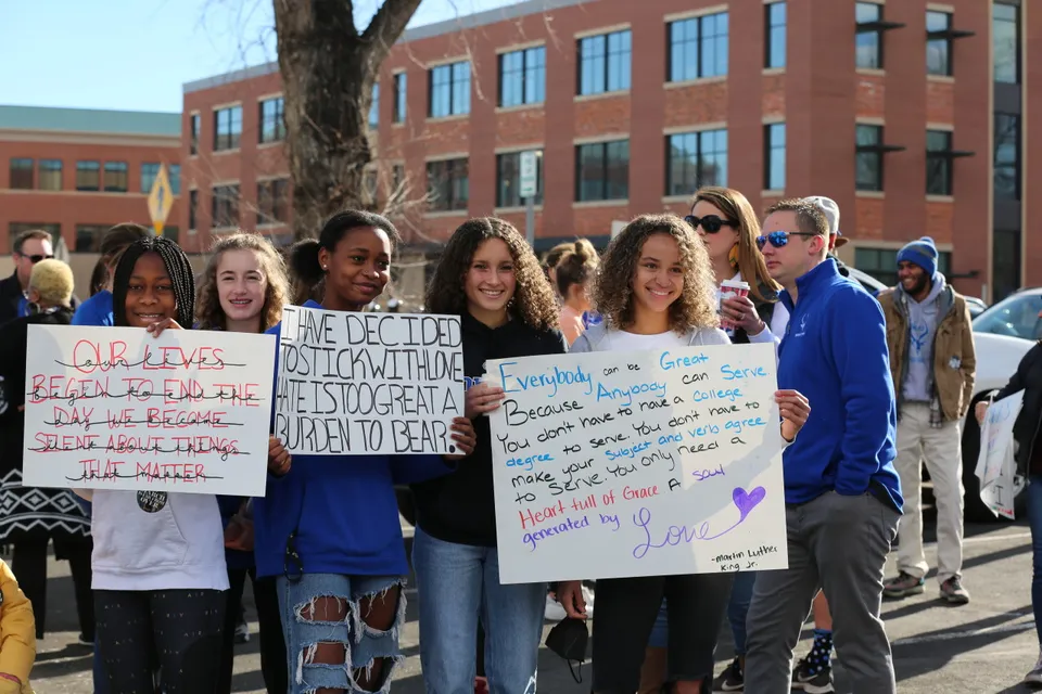 Un gruppo di giovani tiene cartelli con messaggi sociali e politici davanti a un edificio in mattoni nel campus del Poudre School District.