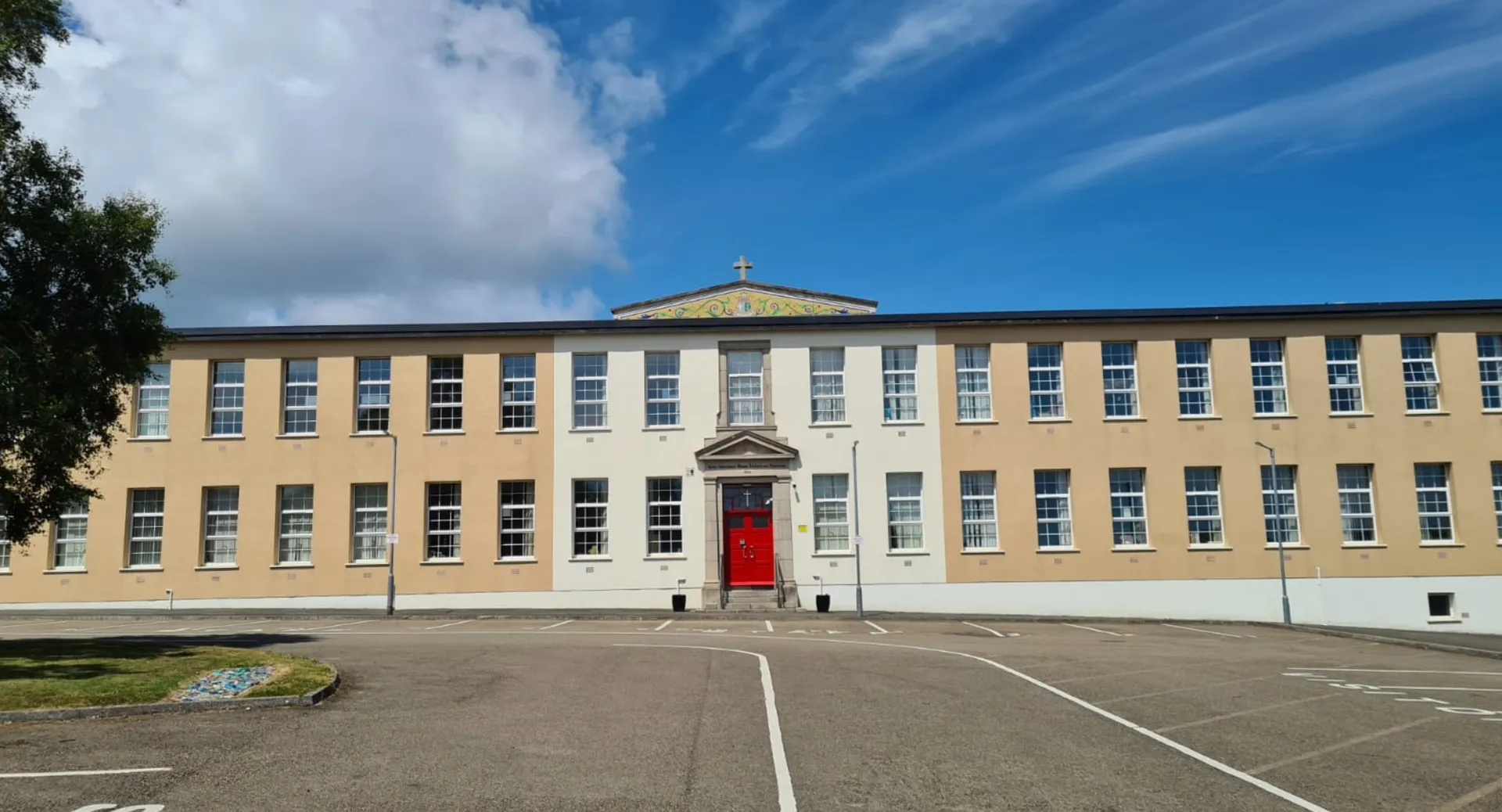 L'edificio scolastico giallo della Mercy Secondary School, con una cupola sulla cima del tetto, è circondato da un piazzale pavimentato.