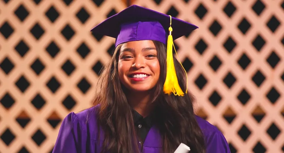 Una diplomanda della Hanalani Schools indossa una toga di diploma viola e sta in piedi davanti a una struttura in legno.