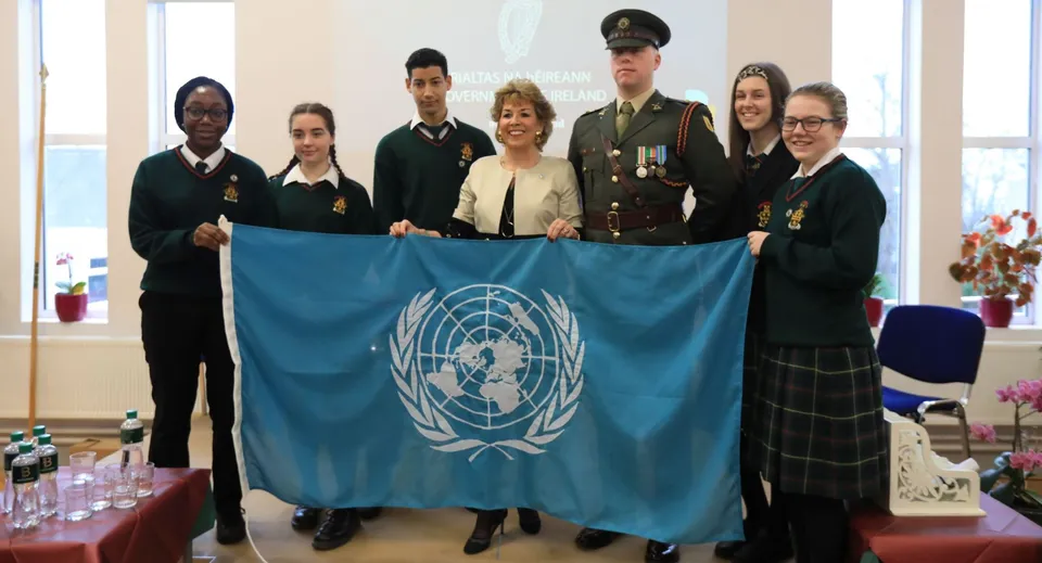 Studenti della Drogheda Grammar School e persone in uniforme militare tengono insieme una bandiera delle Nazioni Unite in un ambiente interno.