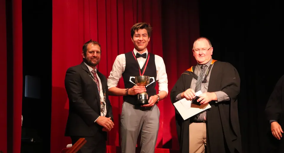 Tre uomini sono sul palco con un sipario rosso, uno di loro tiene una coppa durante una cerimonia di premiazione al Waihi College.