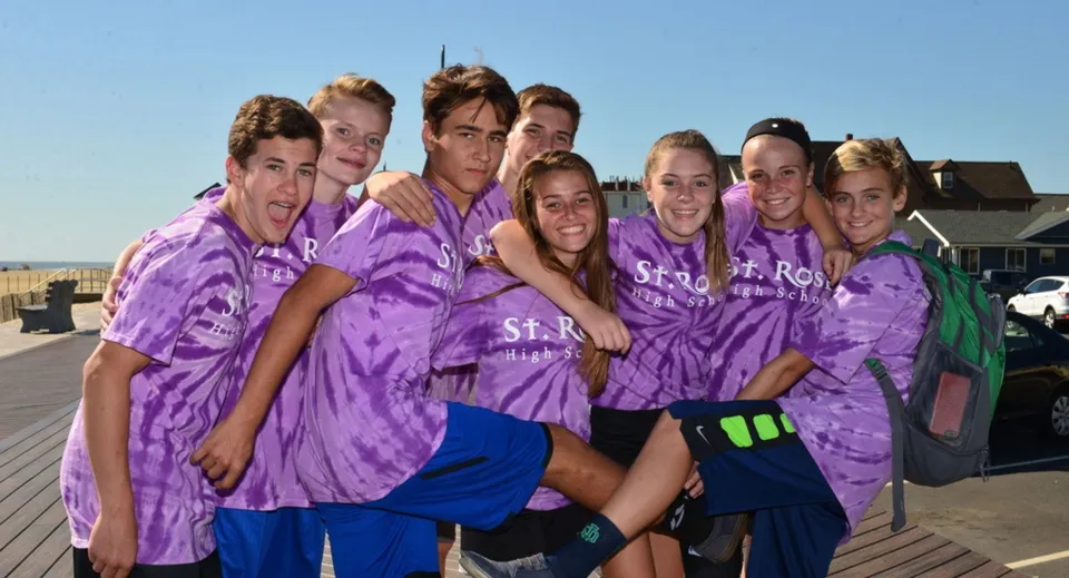Un gruppo di studenti della St. Rose High School indossa t-shirt tie-dye viola e si trova insieme su una terrazza in legno.