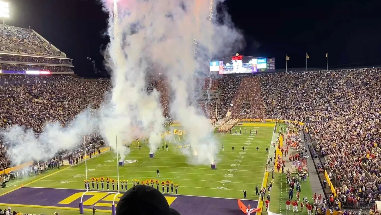 Estadio de fútbol americano de noche con fuegos artificiales, columnas de humo sobre el campo y animadoras ante las gradas llenas