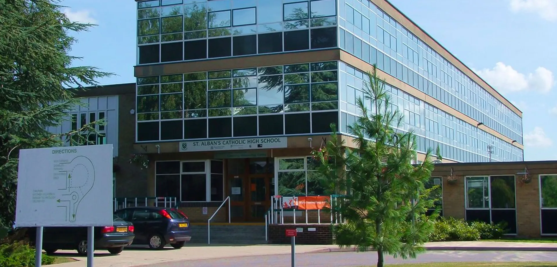 L'edificio moderno in vetro e acciaio della St. Alban's Catholic High School si erge davanti a un cielo azzurro con alberi e un'auto in primo piano.