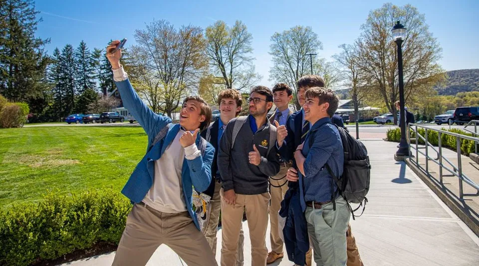 Un gruppo di studenti si trova all'esterno della Trinity-Pawling School in una giornata di sole, con un prato e alberi sullo sfondo.
