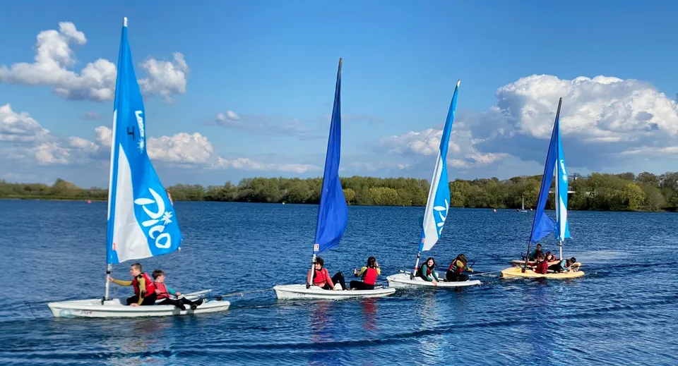 Studenti della Theale Green School vanno in barca a vela con piccole imbarcazioni su un lago calmo circondato da un paesaggio verde.