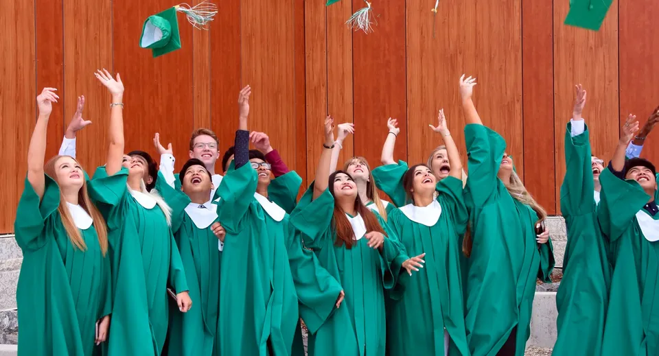 I diplomati e le diplomate del Kildonan East Collegiate in toga verde festeggiano davanti a una parete di legno.