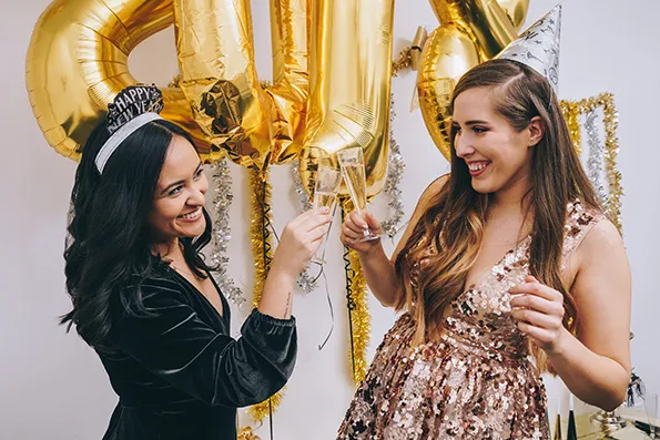 Dos chicas sonrientes brindan con copas frente a globos dorados en una celebración festiva