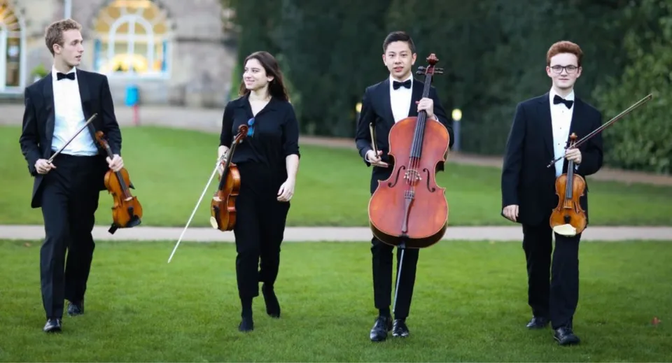 Quattro musicisti della Wells Cathedral School si trovano su un prato con i loro strumenti ad arco davanti a un edificio.