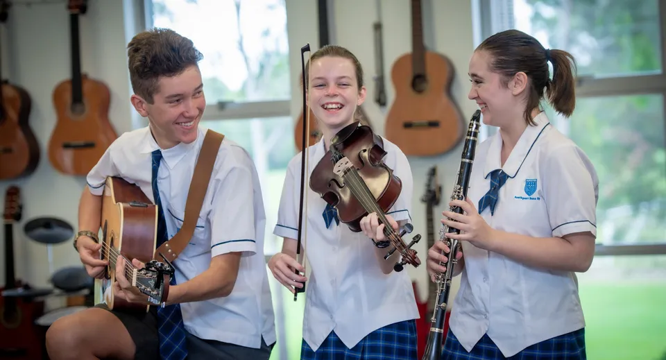 Tre studenti della Southport State High School suonano la chitarra in un'aula con chitarre appese alle pareti.