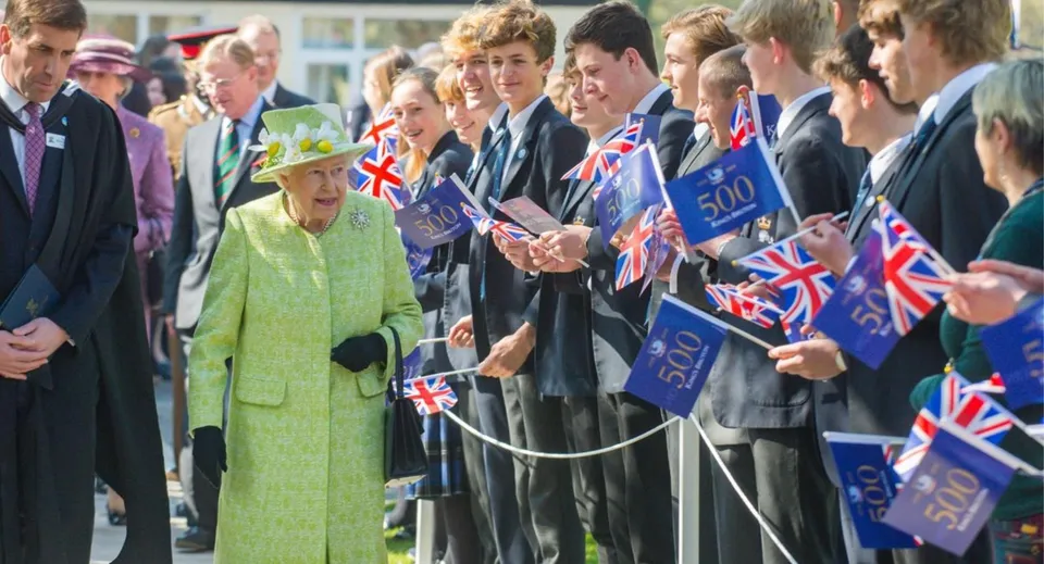 Studenti della King's Bruton accolgono una donna in cappotto verde con bandiere e striscioni britannici.
