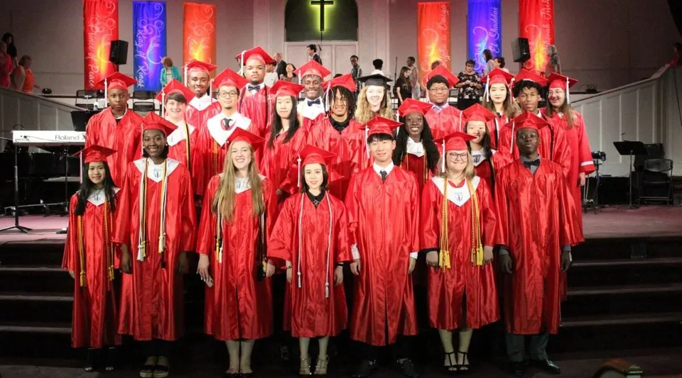 Un gruppo di diplomati della Northside Christian Academy indossa toge rosse in una chiesa con una croce sullo sfondo.
