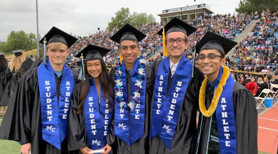 Un gruppo di neolaureati in toghe blu e gialle si trova davanti a un vasto pubblico durante la cerimonia di diploma del Ventura County Community College District.