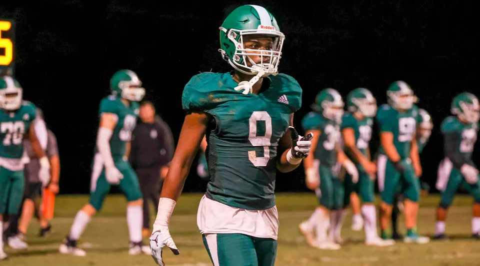 Un giocatore di football della Christ School in maglia verde in campo con i compagni di squadra sullo sfondo.