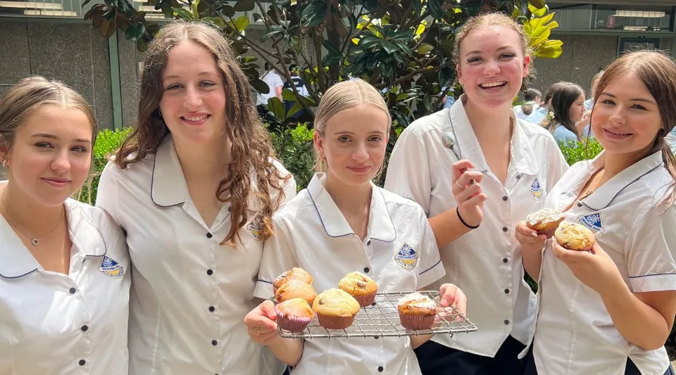 Studentesse della Engadine High School mostrano un vassoio con prodotti da forno appena sfornati su uno sfondo verde.
