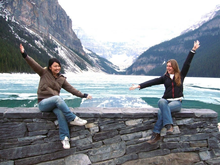 Zwei junge Frauen breiten auf einer Steinmauer vor einem teils zugefrorenen Bergsee die Arme aus