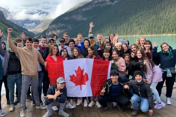 Grupo de jóvenes sosteniendo la bandera de Canadá junto a un lago y montañas