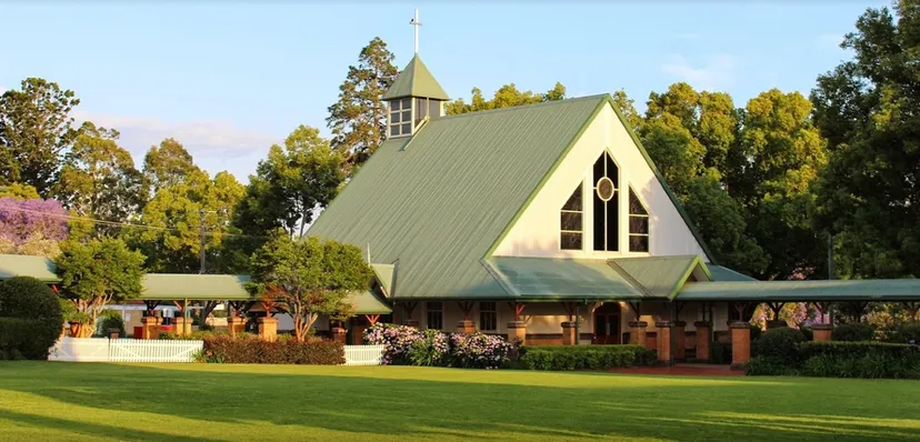 Una chiesa con un tetto a punta e una torre si trova nel campus della Toowoomba Anglican School, circondata da un paesaggio verde e alberi.