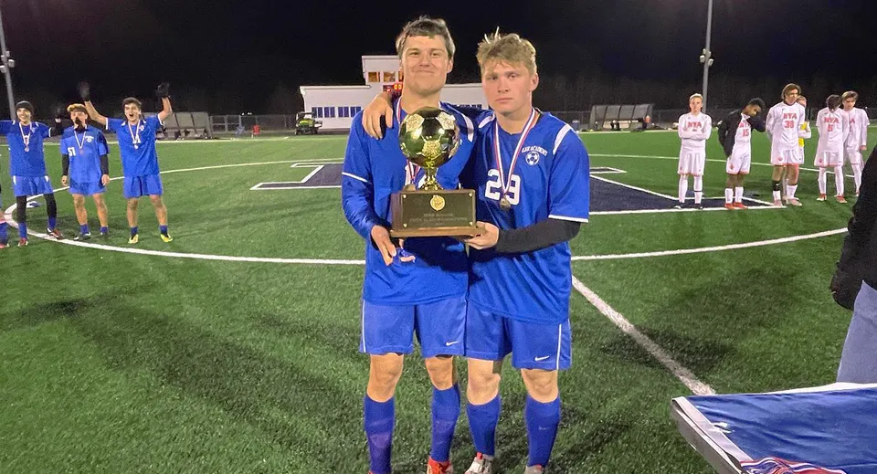 Due calciatori della Lee Academy in divisa blu tengono un trofeo su un campo da gioco di notte.