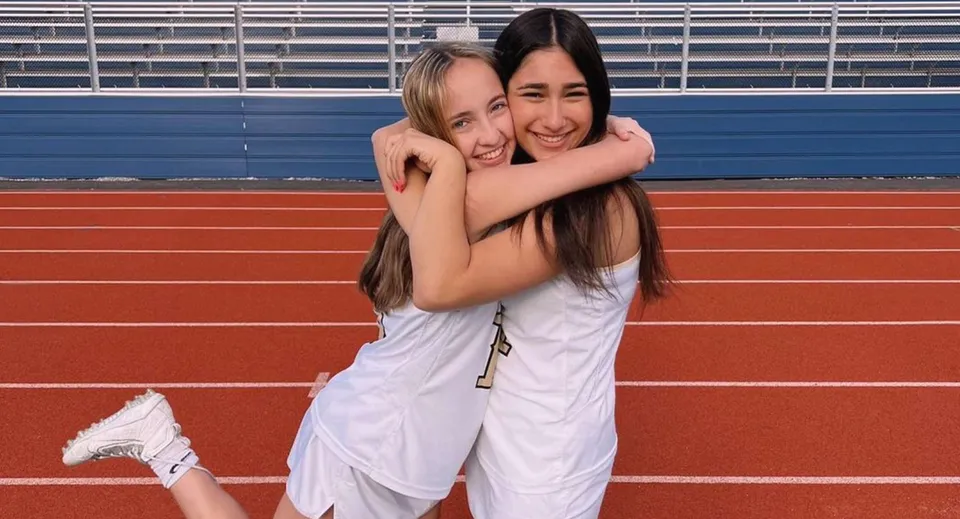Due studentesse del Las Virgenes Unified School District si abbracciano su una pista di atletica rossa davanti agli spalti di uno stadio.