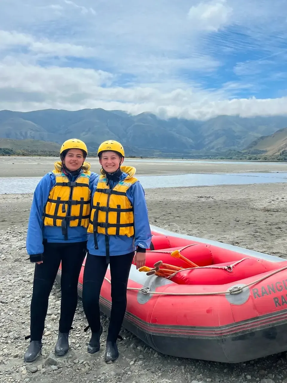 Zwei Mädchen mit Schwimmwesten und Helmen neben einem Raftingboot in Neuseeland