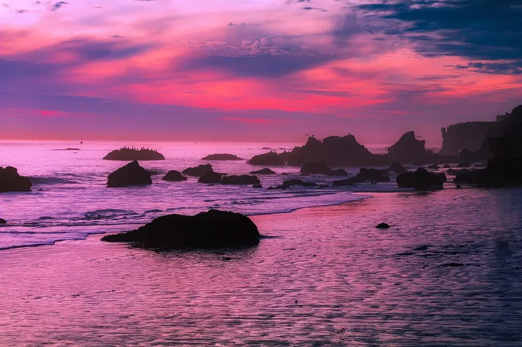 Felsen im Meer vor einer Küste bei pinkfarbenem Sonnenuntergang