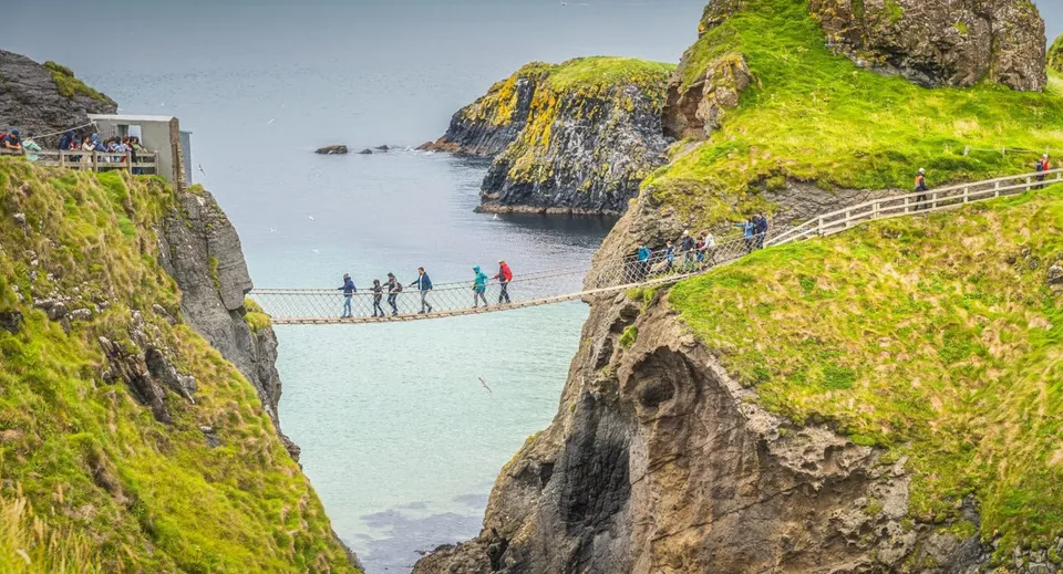 Studenti del Coláiste Mhuire Ballygar attraversano un ponte sospeso sopra un paesaggio costiero con scogliere e acqua.