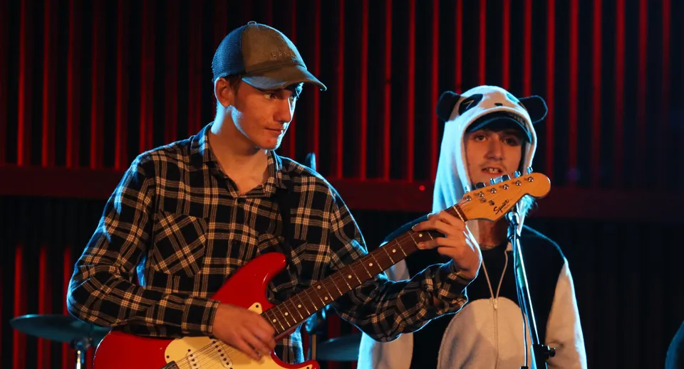 Due studenti dell'Inglewood High School si esibiscono su un palco con tenda rossa, uno di loro suona la chitarra.
