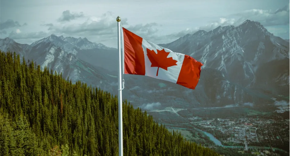 Una grande bandiera canadese sventola davanti a montagne innevate e a una valle boscosa nel campus della A.Y. Jackson Secondary School.