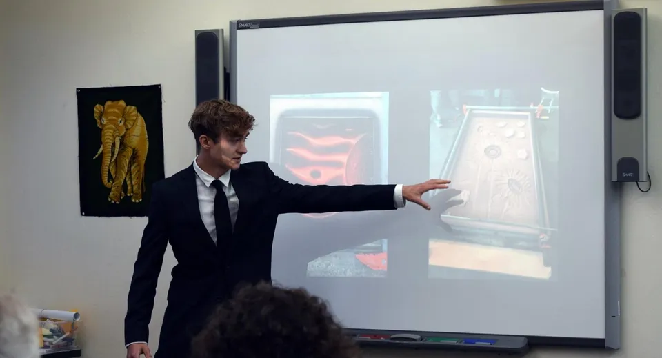 Un uomo in abito elegante sta davanti a un grande schermo di proiezione indicando qualcosa presso la Colorado Rocky Mountain School.