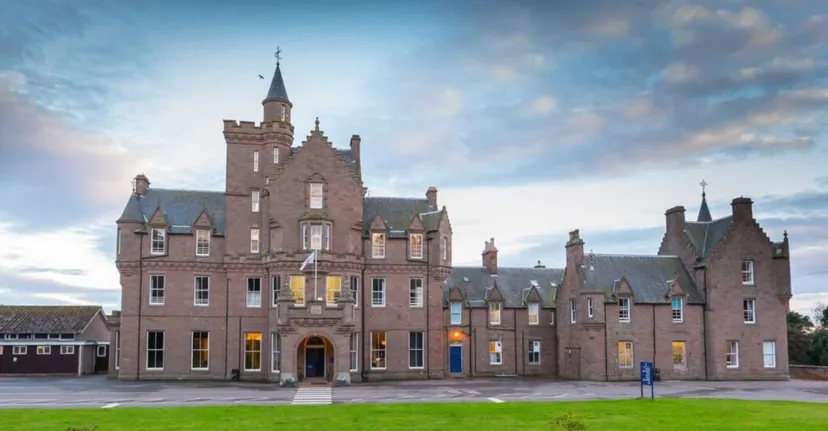 L'imponente edificio simile a un castello della Lathallan School con torri si erge davanti a edifici più piccoli e a un prato sotto un cielo nuvoloso.