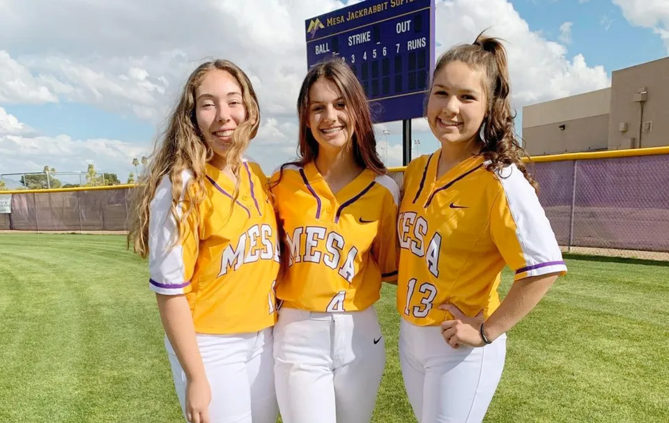 Tre studentesse della Mesa High School in maglie sportive gialle su un campo da baseball con un tabellone sullo sfondo.