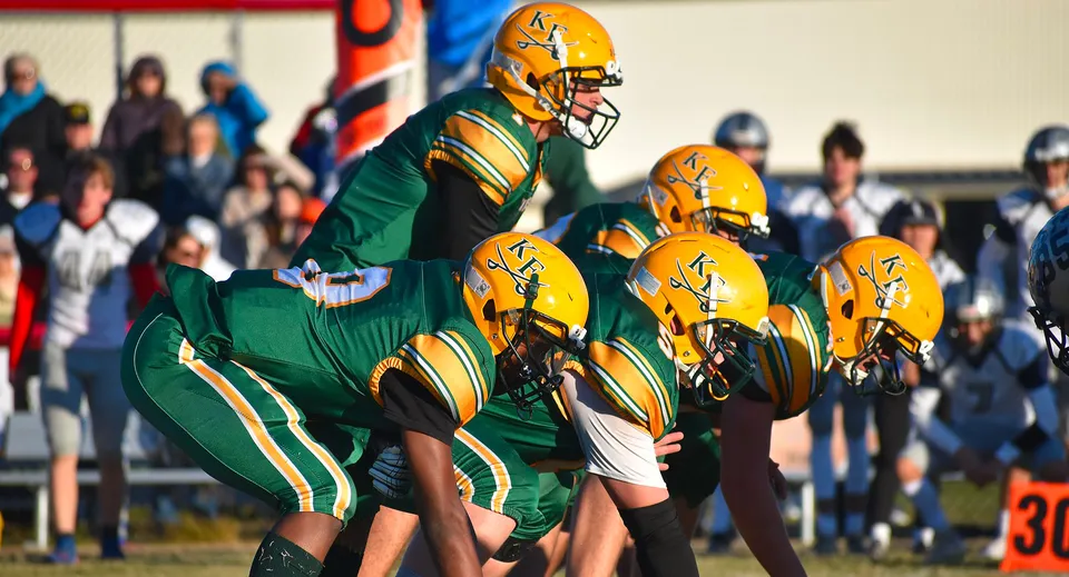 Un gruppo di giocatori di football in maglia verde e gialla si trova sul campo da gioco della Kenston Forest School con spettatori sullo sfondo.