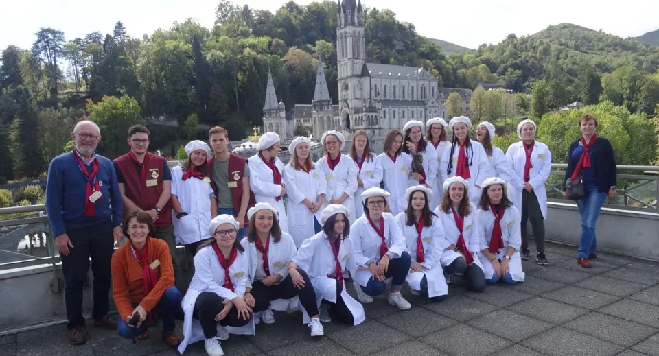Un gruppo di persone in tuniche bianche si trova davanti al grande edificio decorato della Institution Mongazon con alberi e montagne sullo sfondo.