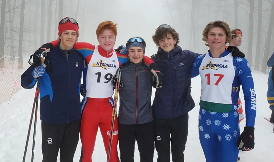 Un gruppo di studenti della Queensbury High School è in piedi con abbigliamento da sport invernali in un paesaggio boschivo innevato.