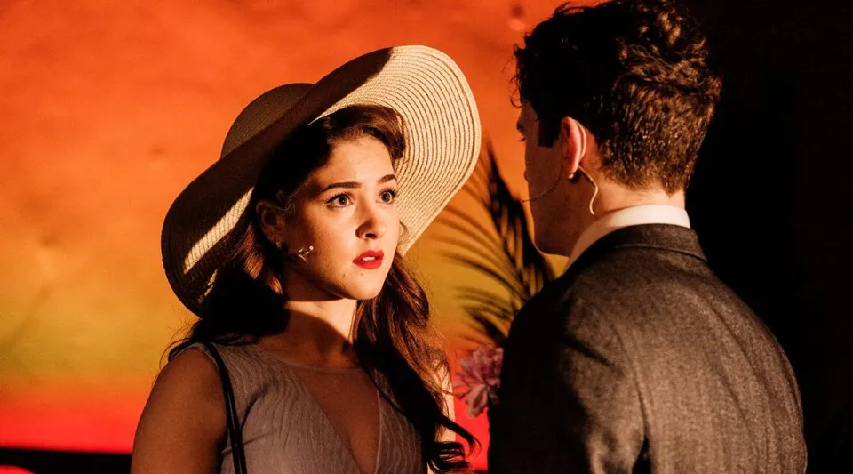 Una donna con un cappello a tesa larga e un uomo in abito elegante sono impegnati in un intenso dialogo davanti a uno sfondo arancione nel campus della Tring Park School for the Performing Arts.