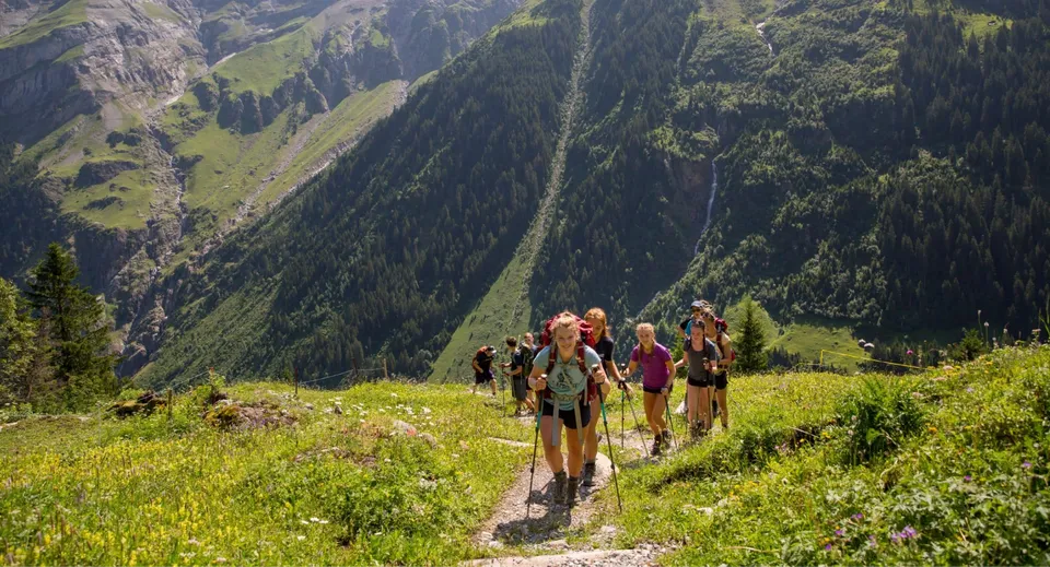 Un gruppo di studenti del Bradfield College fa un'escursione attraverso un paesaggio montano verde con un fitto bosco sullo sfondo.