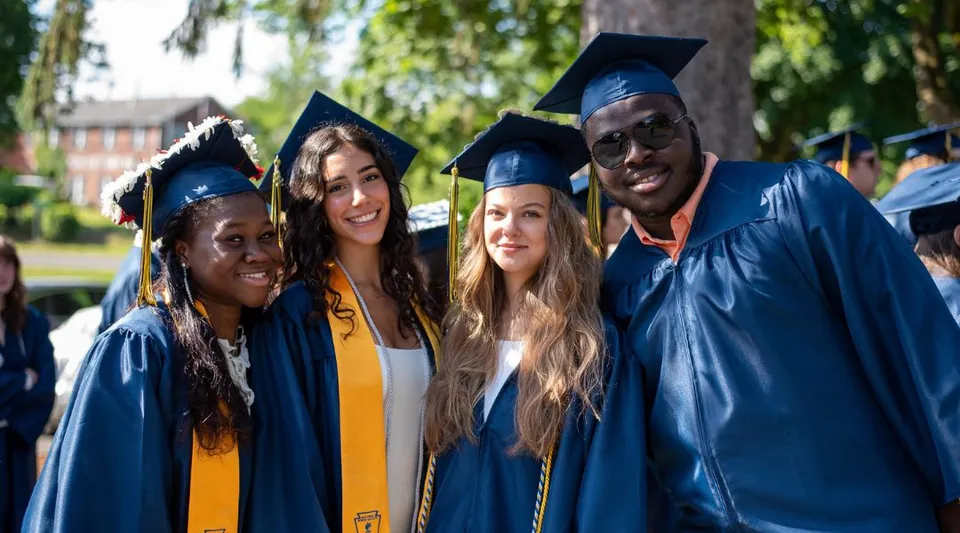 Un gruppo di diplomati e diplomate della Cheshire Academy in toghe blu si trova insieme in un parco con alberi ed edifici sullo sfondo.
