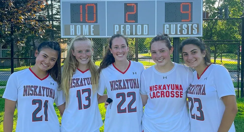 Un gruppo di cinque studentesse della Niskayuna High School davanti a un tabellone segnapunti durante una partita di lacrosse sul campo sportivo.