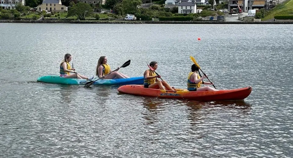 Studenti della Presentation Secondary School (Cork) pagaiano su kayak gonfiabili su uno specchio d'acqua con la città sullo sfondo.