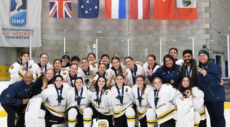 Un gruppo di giocatrici di hockey su ghiaccio della Applecross Senior High School posa davanti a un'arena con bandiere internazionali.