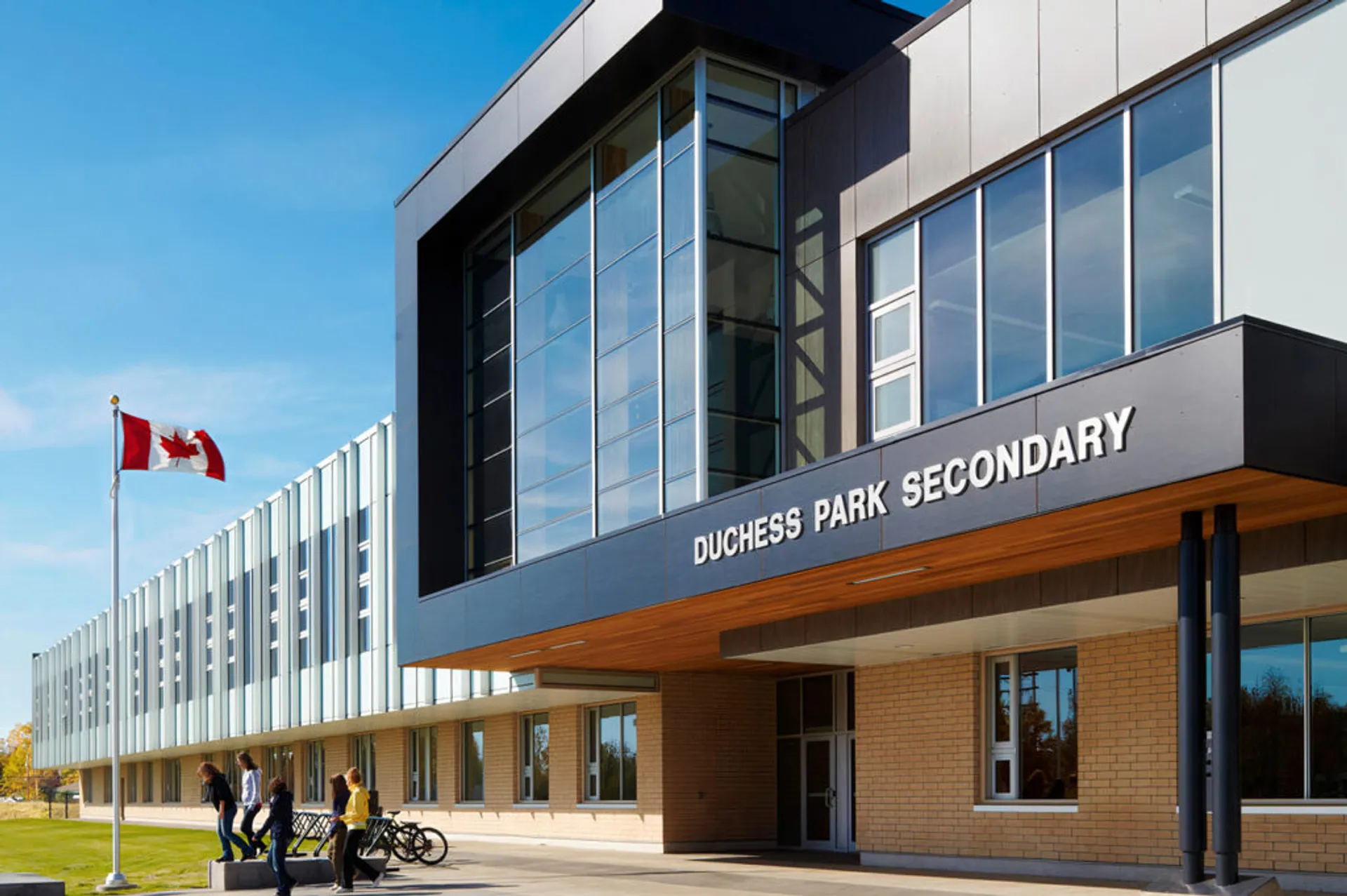 L'edificio moderno a più piani della Duchess Park Secondary School con ampie vetrate sotto un cielo blu e una bandiera canadese in primo piano.