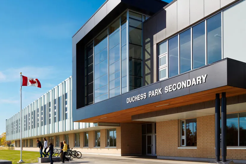 L'edificio moderno a più piani della Duchess Park Secondary School con ampie vetrate sotto un cielo blu e una bandiera canadese in primo piano.