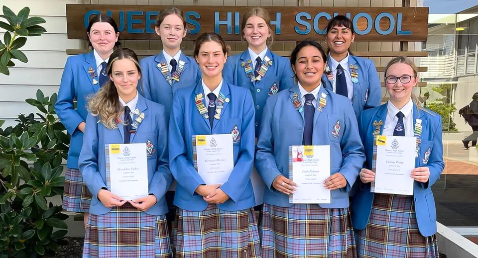 Studentesse e studenti della Queens High School ricevono i loro certificati davanti all'edificio scolastico.