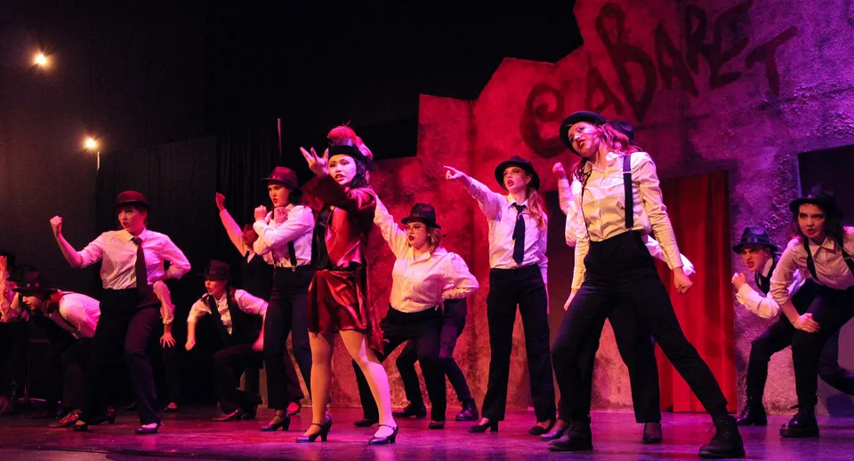 Un gruppo di studenti della Royal Bay Secondary School si esibisce in una vivace performance di danza su un palco con illuminazione rossa e viola.
