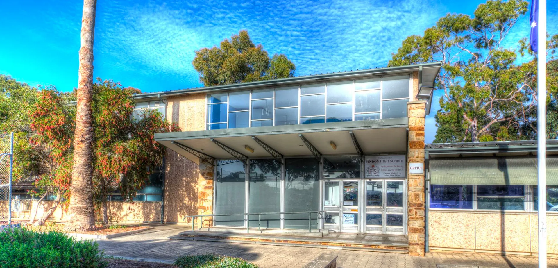 Il moderno edificio in vetro della Findon High School è circondato da una verde vegetazione sotto un cielo azzurro limpido.