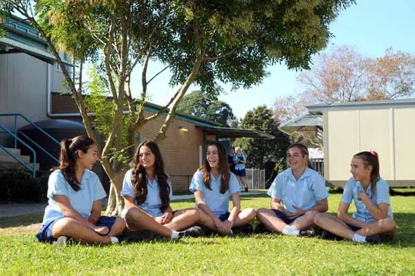 Un grupo de jóvenes alumnas está sentado en el césped en el recinto del Mackellar Girls Campus, rodeado de árboles y edificios.