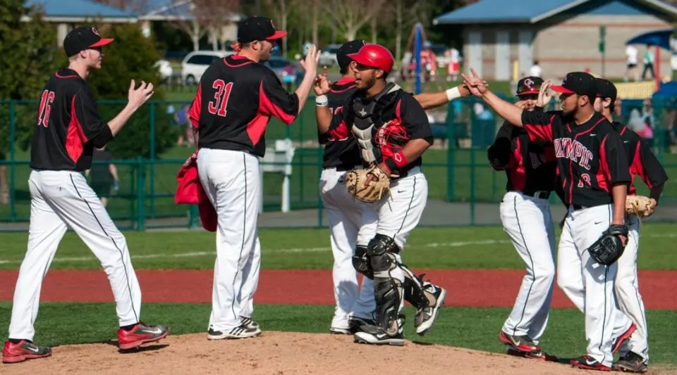 Un gruppo di giocatori di baseball dell'Olympic College festeggia sul campo con edificio e alberi sullo sfondo.