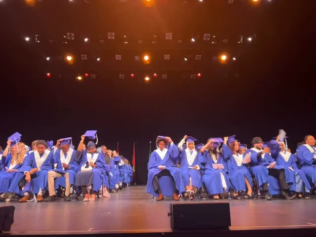 Un grupo de graduados de la École secondaire publique Gisèle-Lalonde está de pie con togas azules sobre un escenario con fondo iluminado.